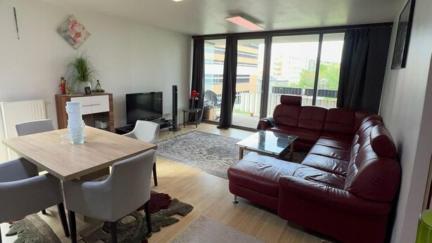 Uma sala de estar moderna com área de jantar, um sofá de couro vermelho, uma TV e uma janela grande com vista para edifícios.