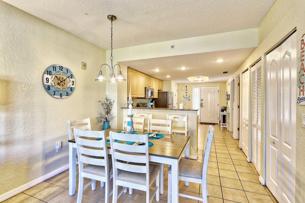 A dining area with a wooden table and chairs, a clock on the wall, and a kitchen in the background. - Sunset Vistas 209n - Latitude Adjustment (Treasure Island)