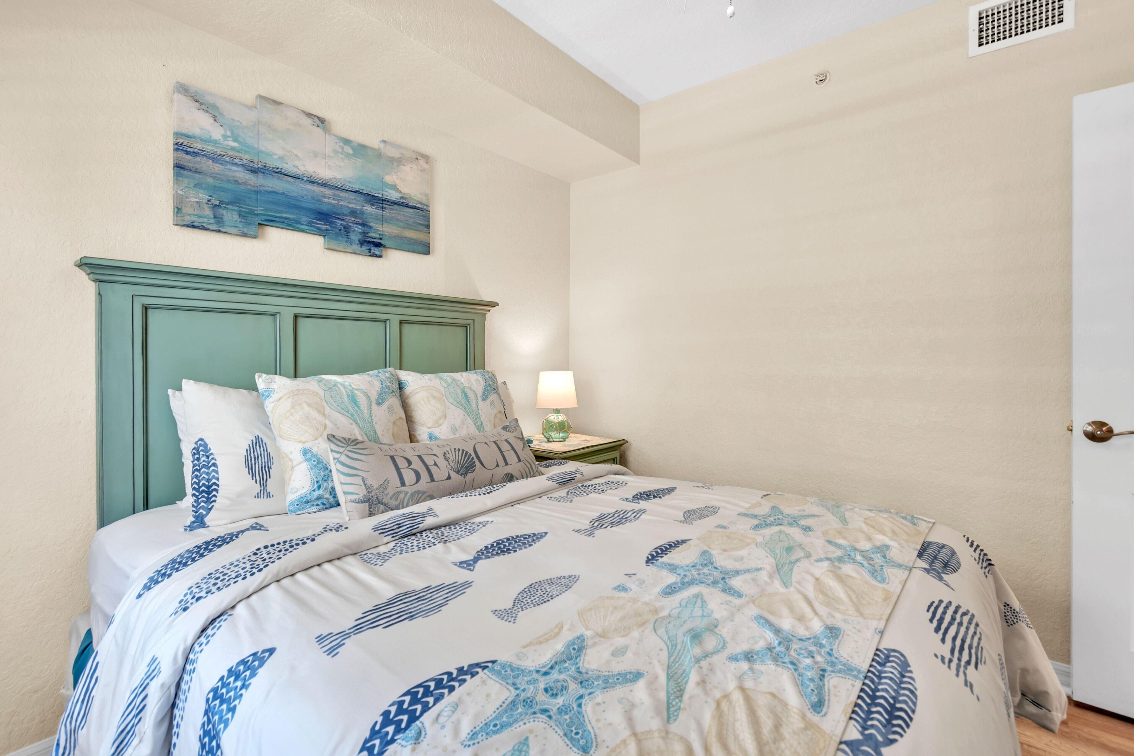 Quarto com cama com colcha de tema náutico, cabeceira verde-água e pintura pendurada na parede retratando uma paisagem marítima.