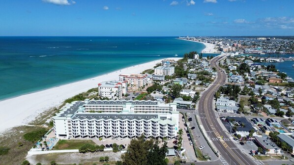 Vista aérea de una ciudad costera con un hotel destacado de varios pisos y balcones, una playa y una calle con mucho tráfico.
