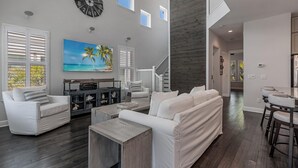 A spacious living room with a large clock, a painting of a beach, and a staircase leading to an upper level.