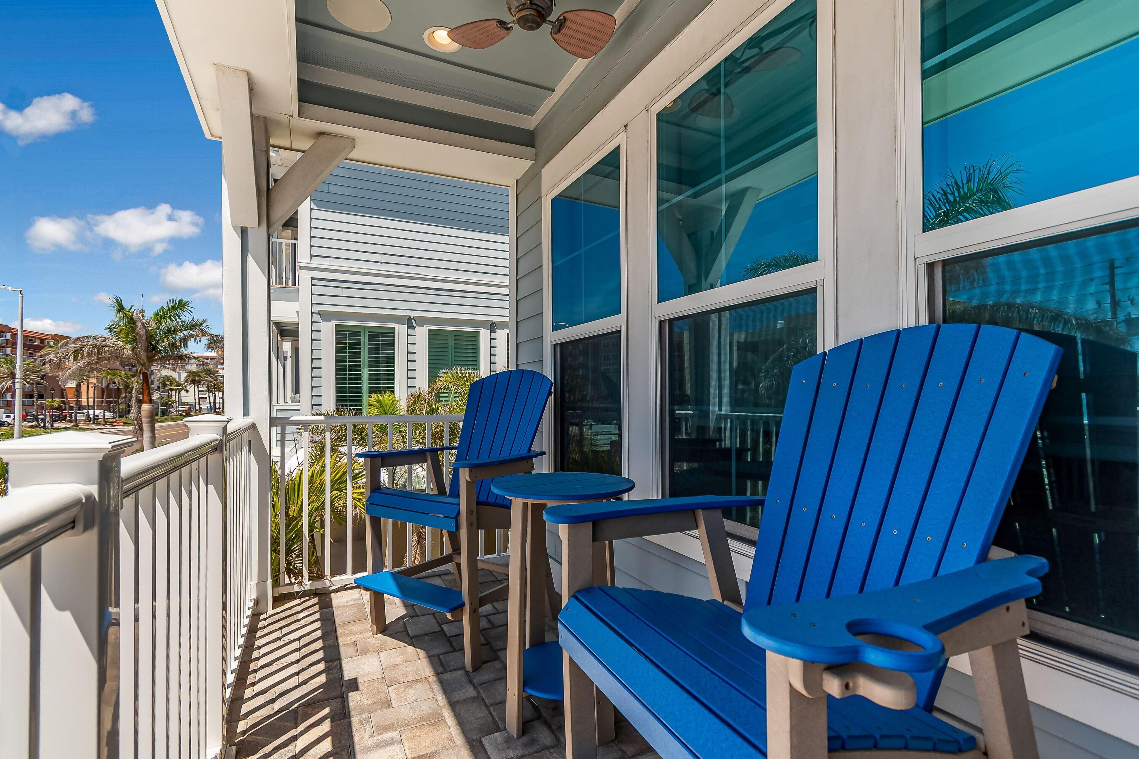 A covered outdoor area with blue Adirondack chairs and tables, white railings, and large windows with reflections of palm trees.