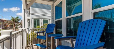 A covered outdoor area with blue Adirondack chairs and tables, white railings, and large windows with reflections of palm trees.