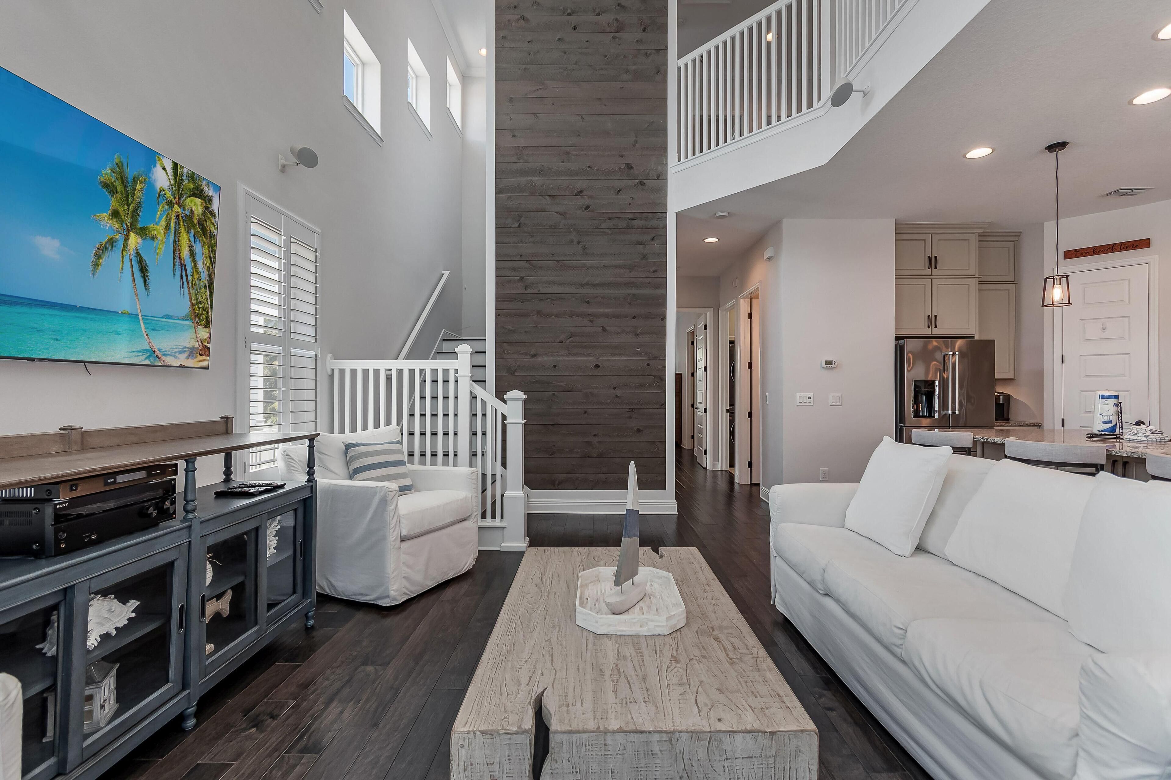Un salon spacieux avec un canapé blanc, une table basse et un grand tableau représentant une scène de plage. Un escalier avec une rampe mène à l’étage supérieur.