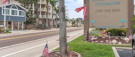 Señal de Sea Club Condominiums en 19725 Gulf Blvd, con banderas estadounidenses y palmeras.