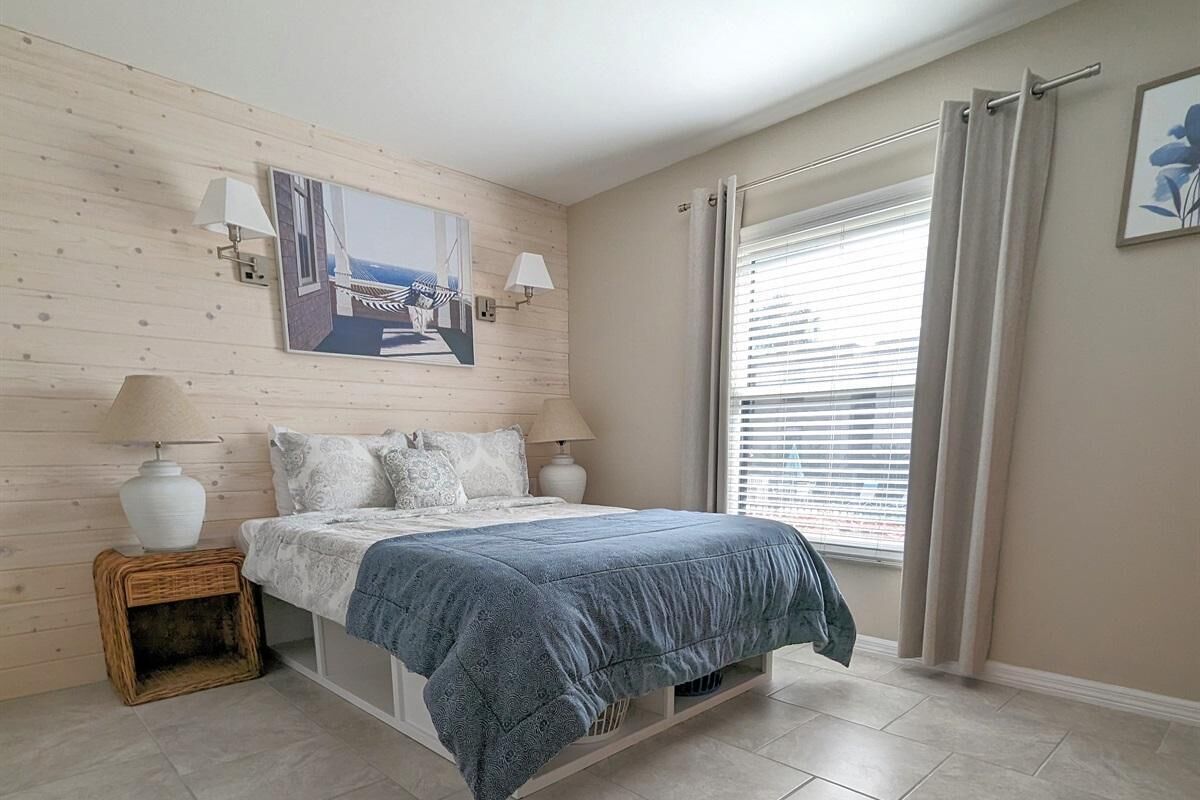 Ein Schlafzimmer mit einem Bett, zwei Nachttischlampen, einem Bild an der Wand, einem Fenster mit Vorhängen und einem kleinen Korbwäscheschrank.