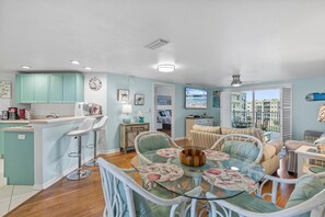 A spacious living area with a glass dining table, white wicker chairs, and a view of the city through large windows. - Oceanwalk 1-502 (New Smyrna Beach)