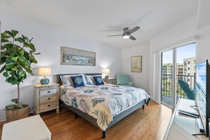 A bedroom with a bed, nightstands, a ceiling fan, a large window, and a balcony. - Oceanwalk 1-502 (New Smyrna Beach)
