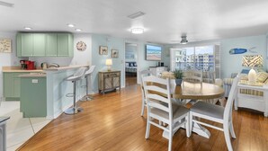A spacious kitchen with green cabinets, a dining area with white chairs, and a view of buildings through the open door.