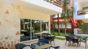 Une terrasse extérieure avec des tables et des chaises, une porte vitrée donnant sur un espace intérieur, et un bâtiment rouge en arrière-plan.