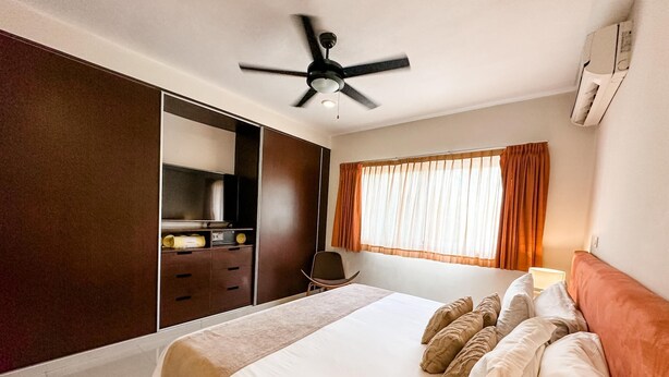 Une chambre à coucher comprenant un lit, un ventilateur de plafond, une télévision, une armoire et une fenêtre avec des rideaux.