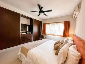 Une chambre à coucher comprenant un lit, un ventilateur de plafond, une télévision, une armoire et une fenêtre avec des rideaux.
