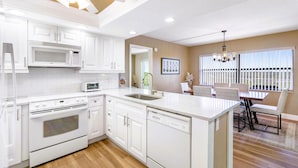 A modern kitchen with white cabinetry, a built-in microwave, and a dishwasher. There is a dining area with a table and chairs, and a ceiling fan above. - Bayshore Yacht & Tennis 806 (Indian Shores)