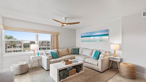 A modern living room with a beige sofa, white coffee table, and a ceiling fan. There are two side tables with lamps and a large window with a view of buildings. - Seaward B503 (New Smyrna Beach)