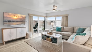 A modern living room with a large TV, a ceiling fan, a sofa, and a view of the outdoors. - Seaward B503 (New Smyrna Beach)