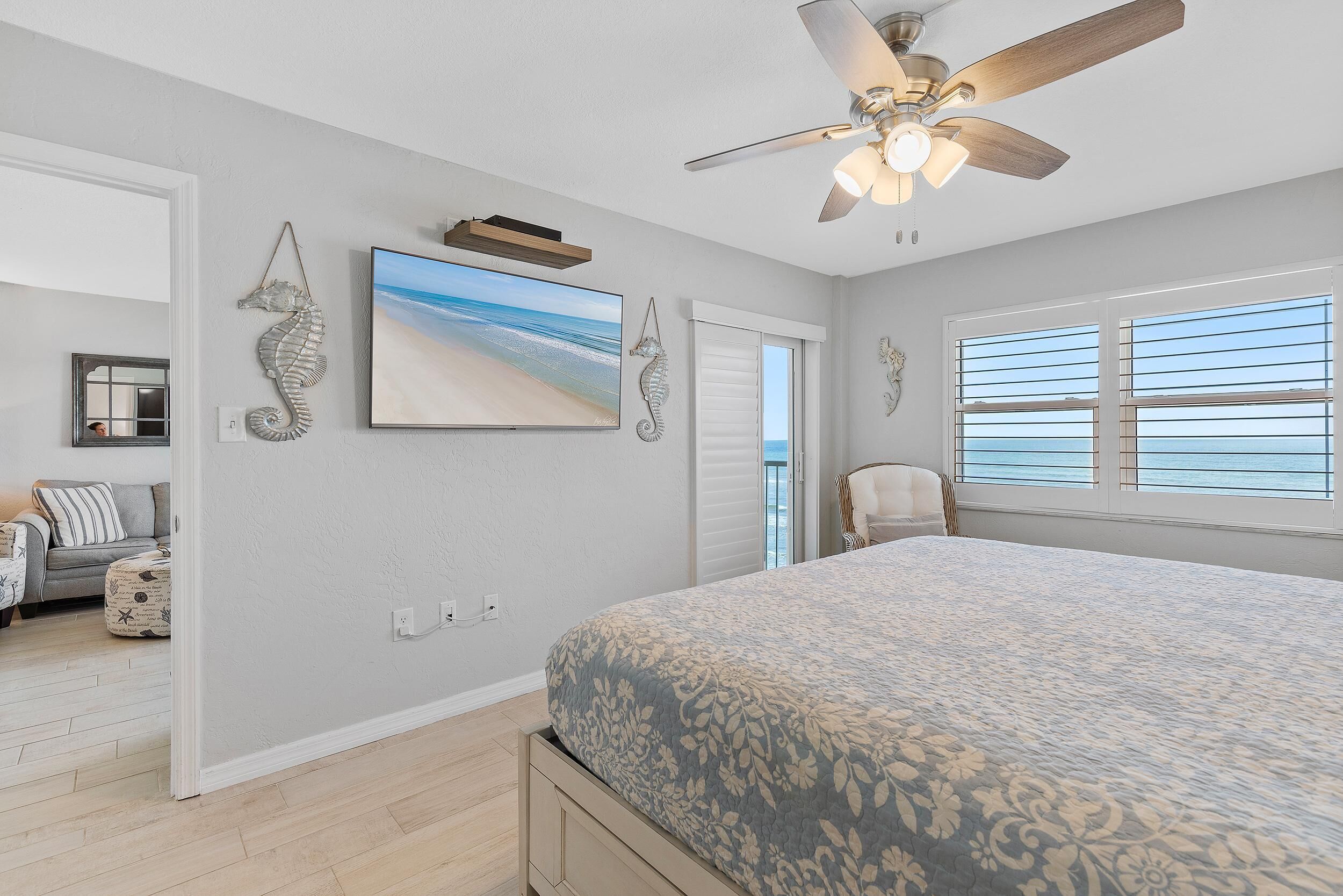 A bedroom with a large bed, a ceiling fan, a beach-themed painting, and a view of the ocean through the window.