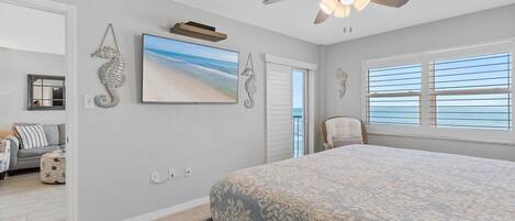 A bedroom with a large bed, a ceiling fan, a beach-themed painting, and a view of the ocean through the window.