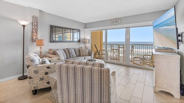 A spacious living room with a large window overlooking a balcony, a sofa set, and a decorative sign on the wall. - Ponce de Leon Towers 804 (New Smyrna Beach)