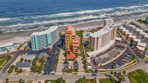 A coastal hotel with multiple buildings, a parking lot, and a beach in the background.