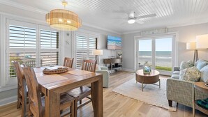 Amplia sala de estar con una mesa de comedor de madera, asientos cómodos, un ventanal con vista a la playa y un ventilador de techo.