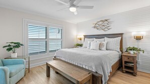 Un dormitorio con cama, mesitas de noche, un banco, ventilador de techo y una ventana con persianas.