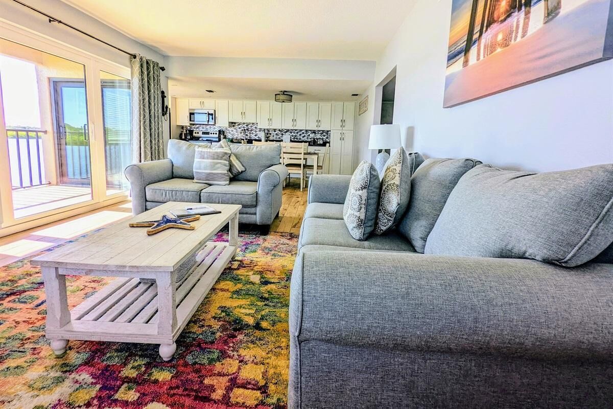 A living room with a grey sofa, a coffee table, and a colorful rug. There is a kitchen in the background with white cabinets and a dining table.