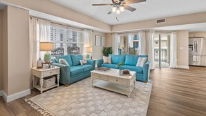 A spacious living room with a ceiling fan, two blue sofas, a coffee table, and a view of the city through large windows. - Oceanwalk 1-401 (New Smyrna Beach)