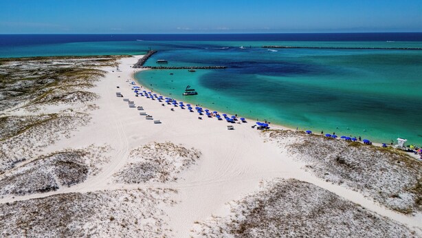 An aerial view of a beach with clear turquoise waters, a sandy shore, and blue umbrellas lined up along the beach. - Coastal Paradise on Holiday Isle - Quiet place, snorkeling cove! Gated Parking! (Destin)