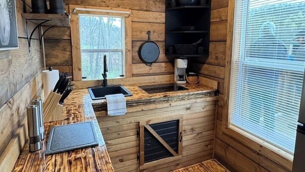 A compact wooden cabin kitchen with a sink, stove, and window. - Glenville’s Rustic Charm (Glenville)