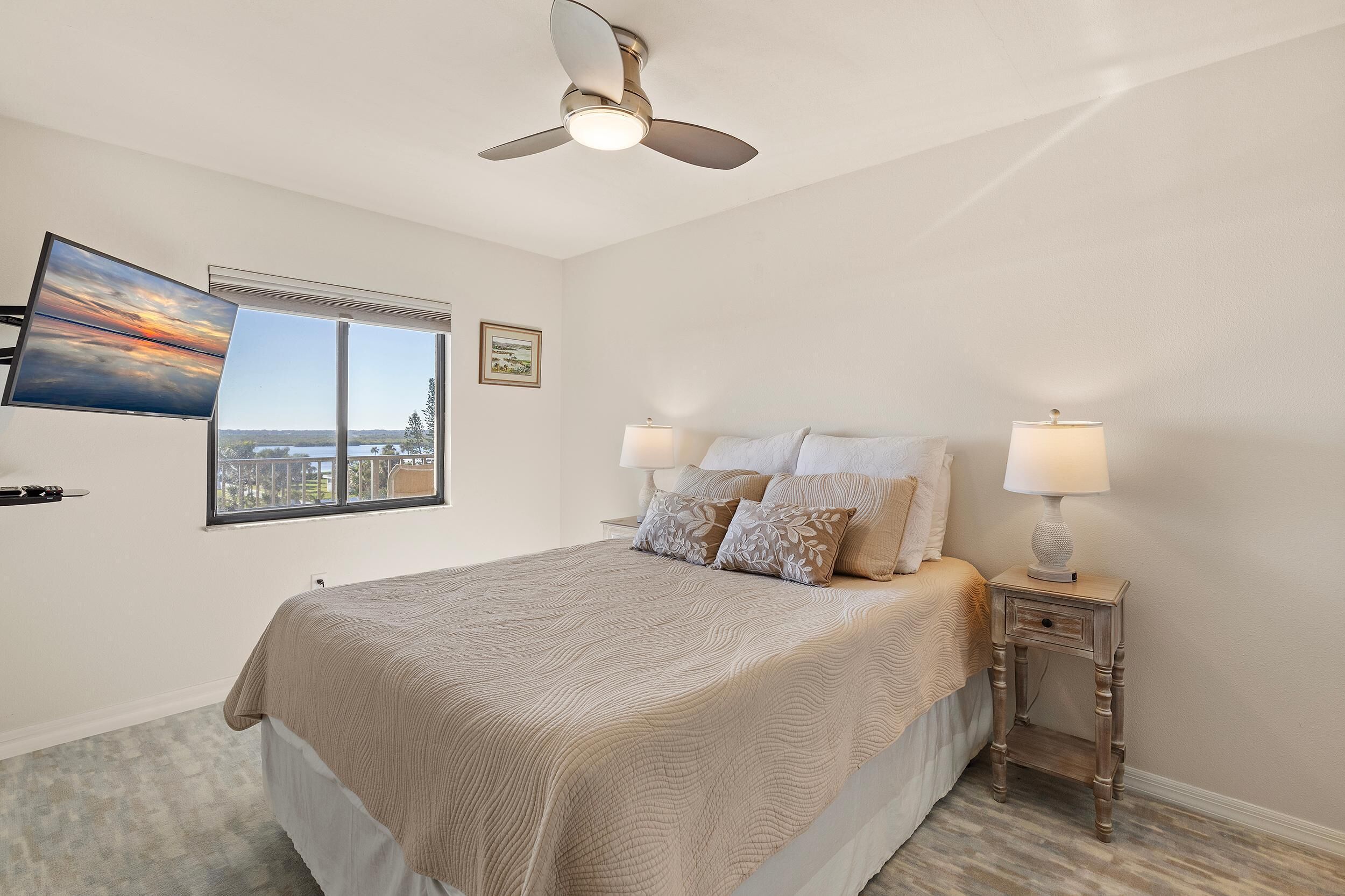 A bedroom with a bed, two bedside tables with lamps, a ceiling fan, a TV mounted on the wall, and a window with a city view.