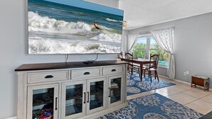 Ein Essbereich mit einem großen Flachbildfernseher, der einen Surfer auf Wellen zeigt, einem Holztisch mit Stühlen und Blick ins Freie durch ein Fenster mit transparenten Vorhängen.