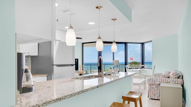 A modern kitchen with a large island, stainless steel appliances, and pendant lighting. There is a view of the ocean through large windows. - Sunbird 309e (Panama City Beach)
