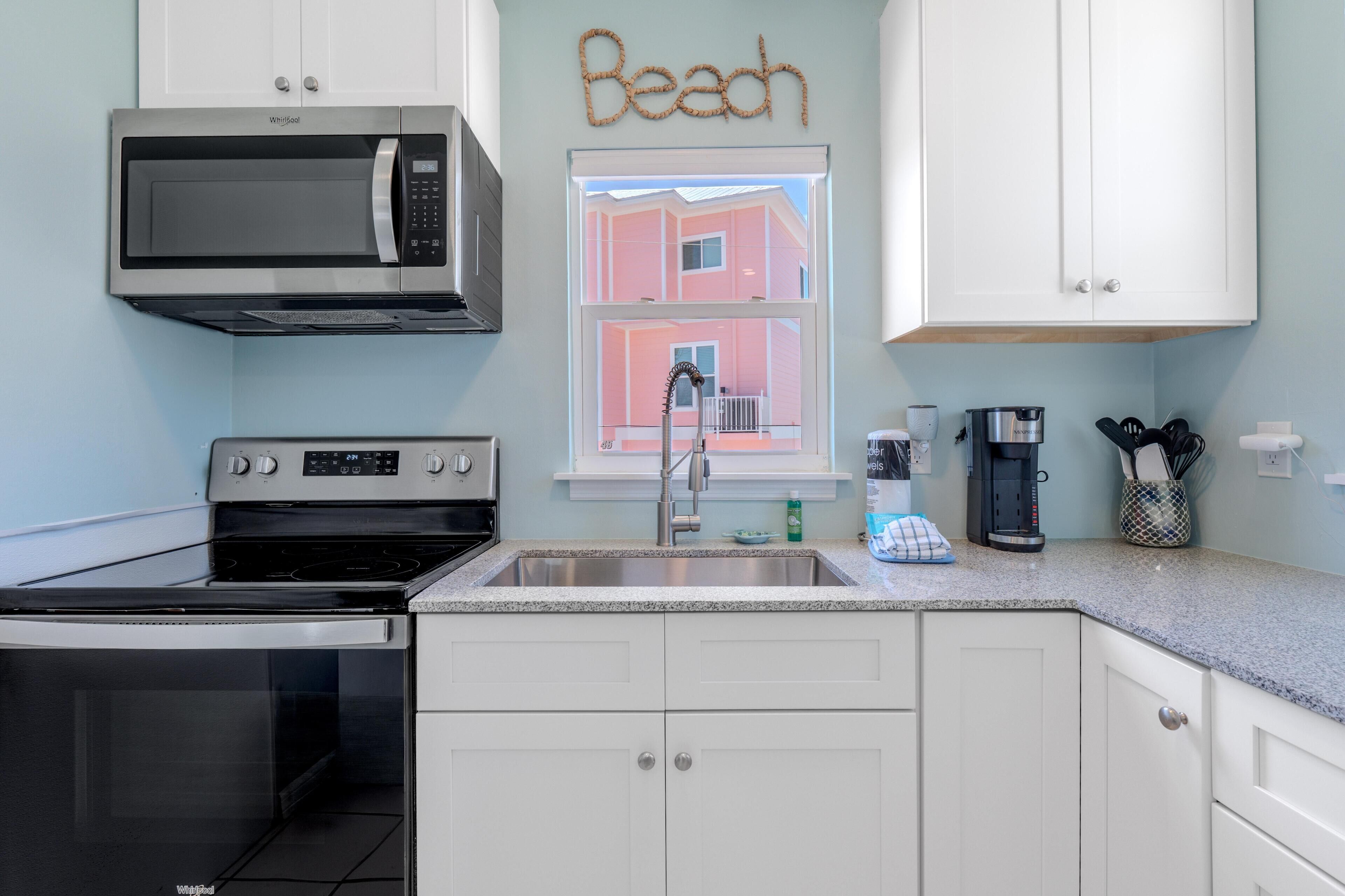 A kitchen with a microwave, stove, sink, and a window with a view of a pink building.