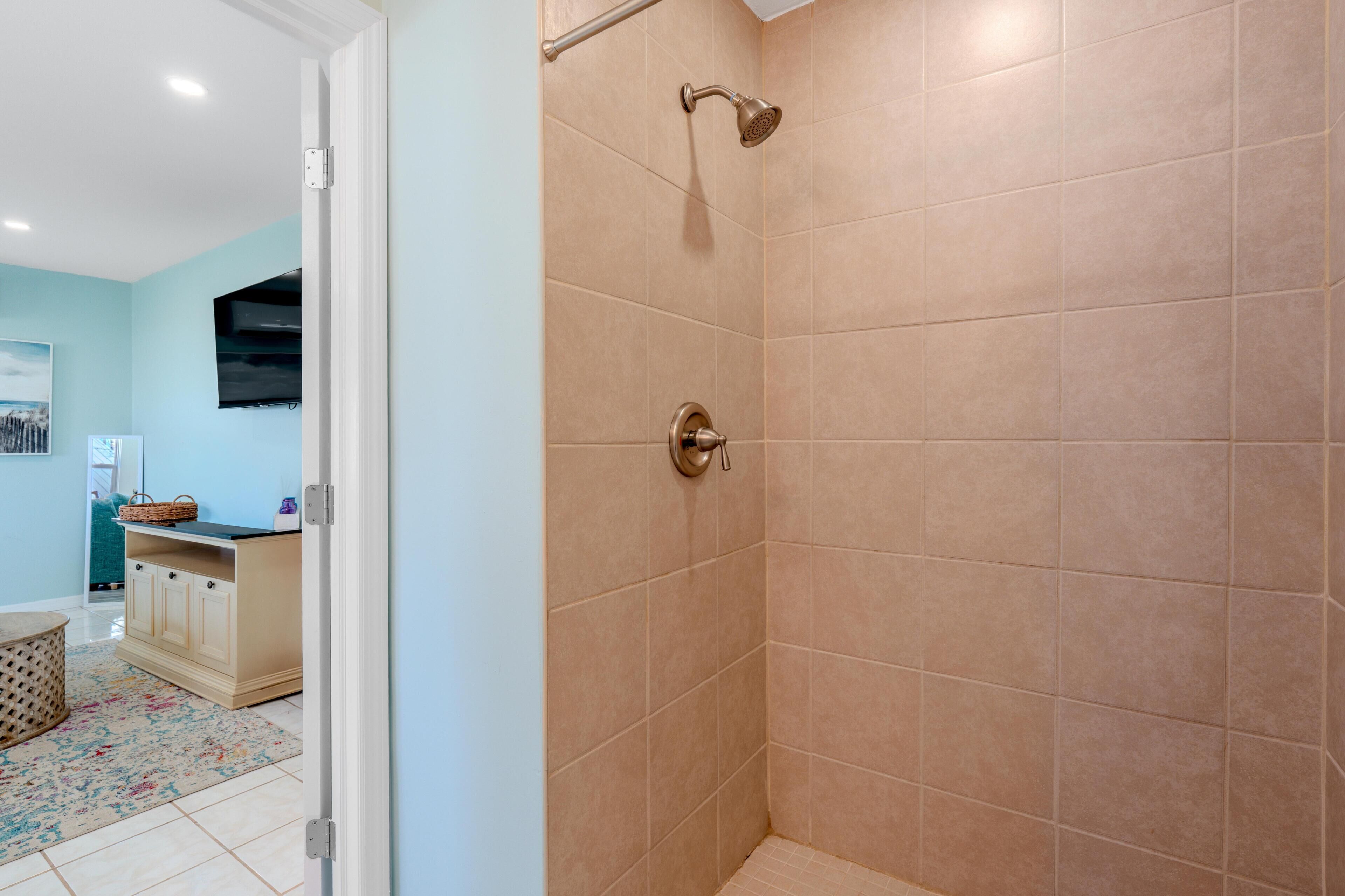 A bathroom with beige tiles, a shower with a handheld showerhead, and a door leading to a room with a blue wall and a wooden cabinet.