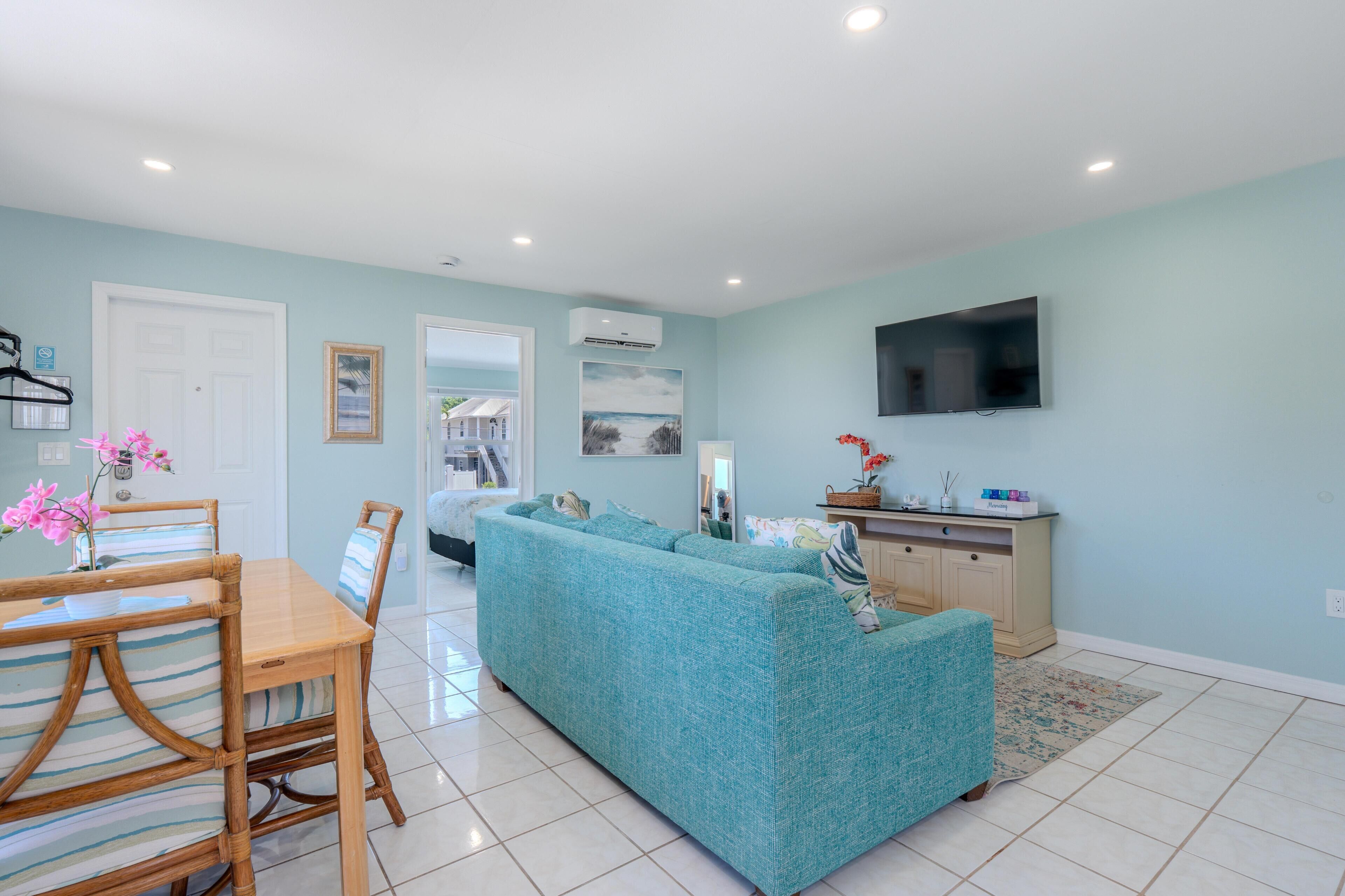 A living room with a sofa, a dining table with chairs, a TV mounted on the wall, and a kitchen area.