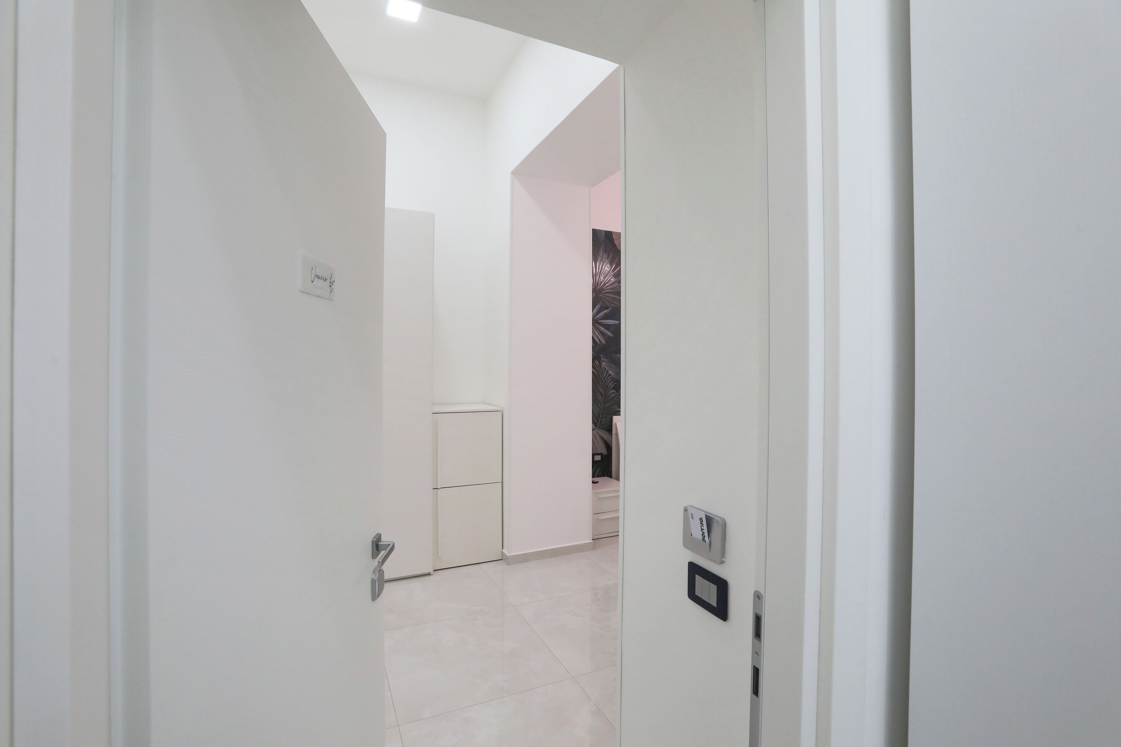 A hallway with a white door, a thermostat, and a small refrigerator in the background.