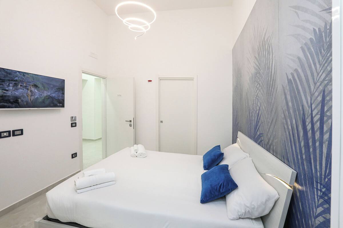 A modern hotel room with a large bed, a flat-screen TV mounted on the wall, a door with a red light, and a wall with a leaf pattern.