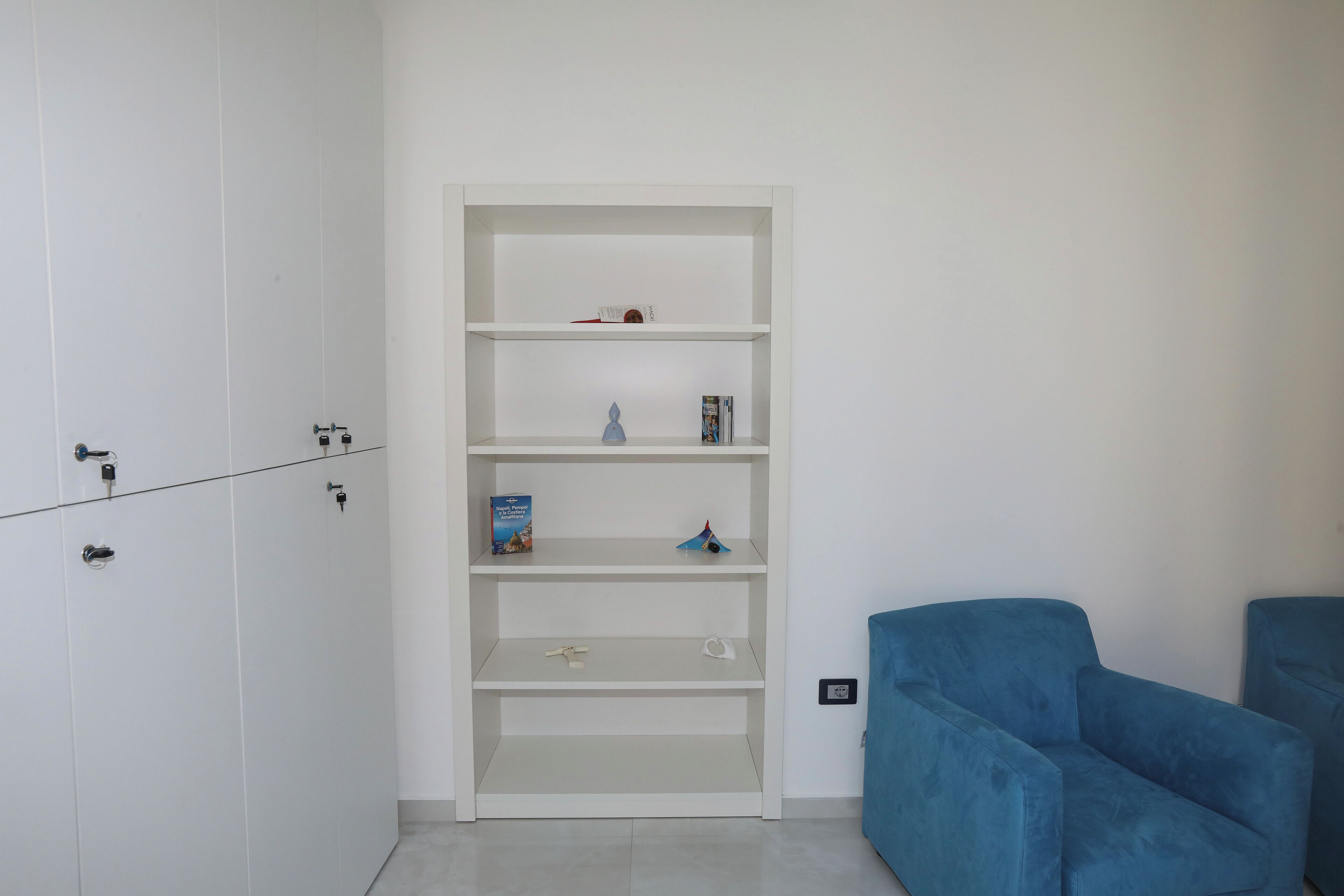 A room with a white wardrobe, a blue sofa, and a white shelving unit with a few items on it.