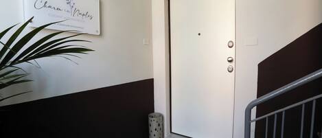 Entrada de hotel com placa indicando "Charm in Naples", tapete de boas-vindas, planta em vaso e porta com fechadura eletrônica.