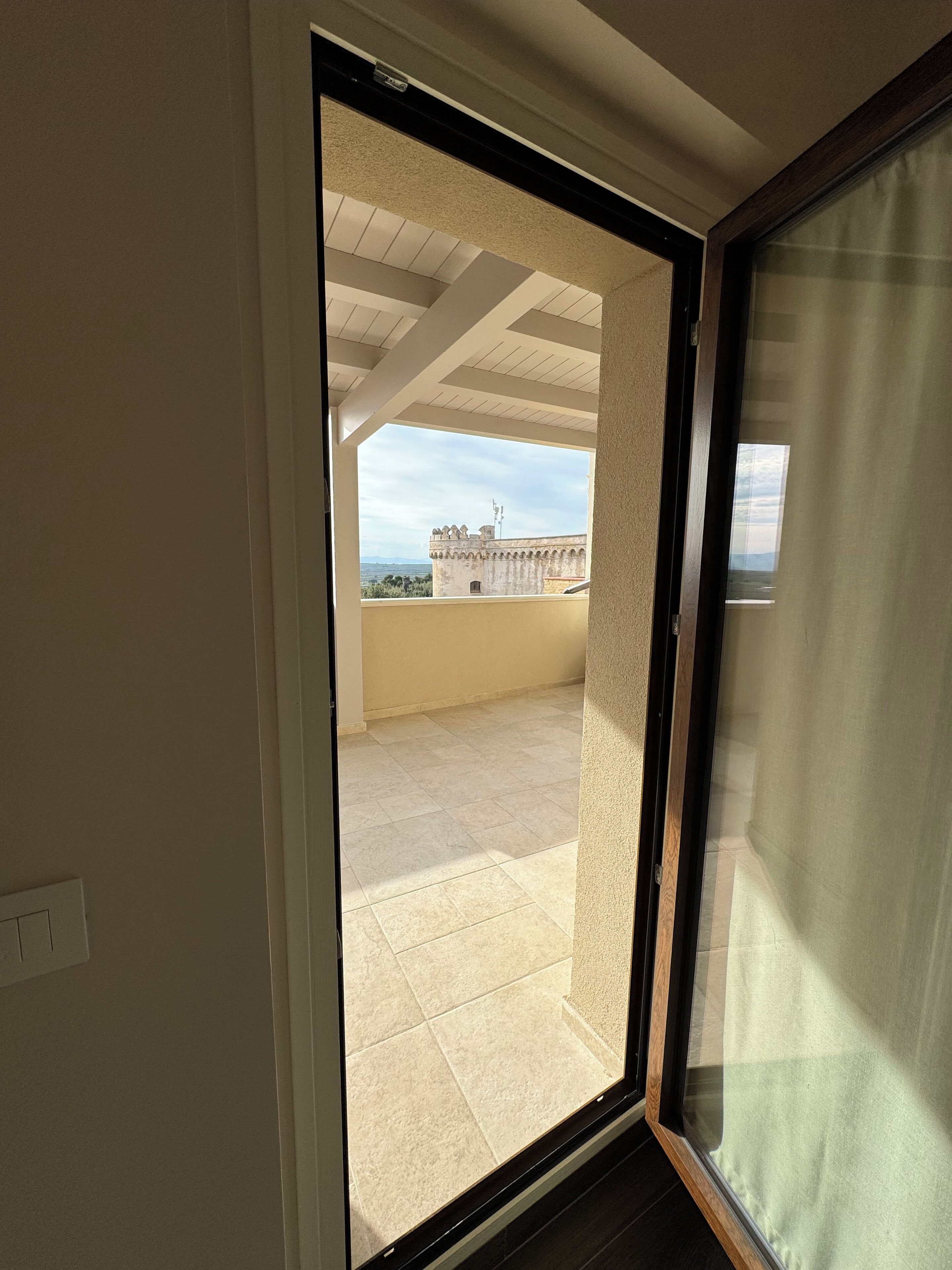 A room with a sliding glass door leading to a balcony with a tiled floor and a view of a building with a tower.