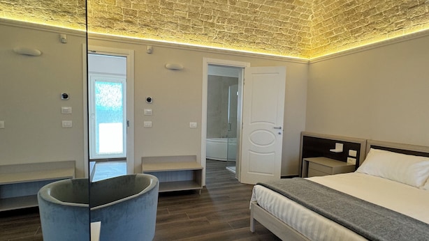 A modern hotel room with a brick-patterned ceiling, a large bed, a bedside table, a chair, and a view of the bathroom through an open door. - B&B PALAZZO CROGHAN (Torremaggiore)