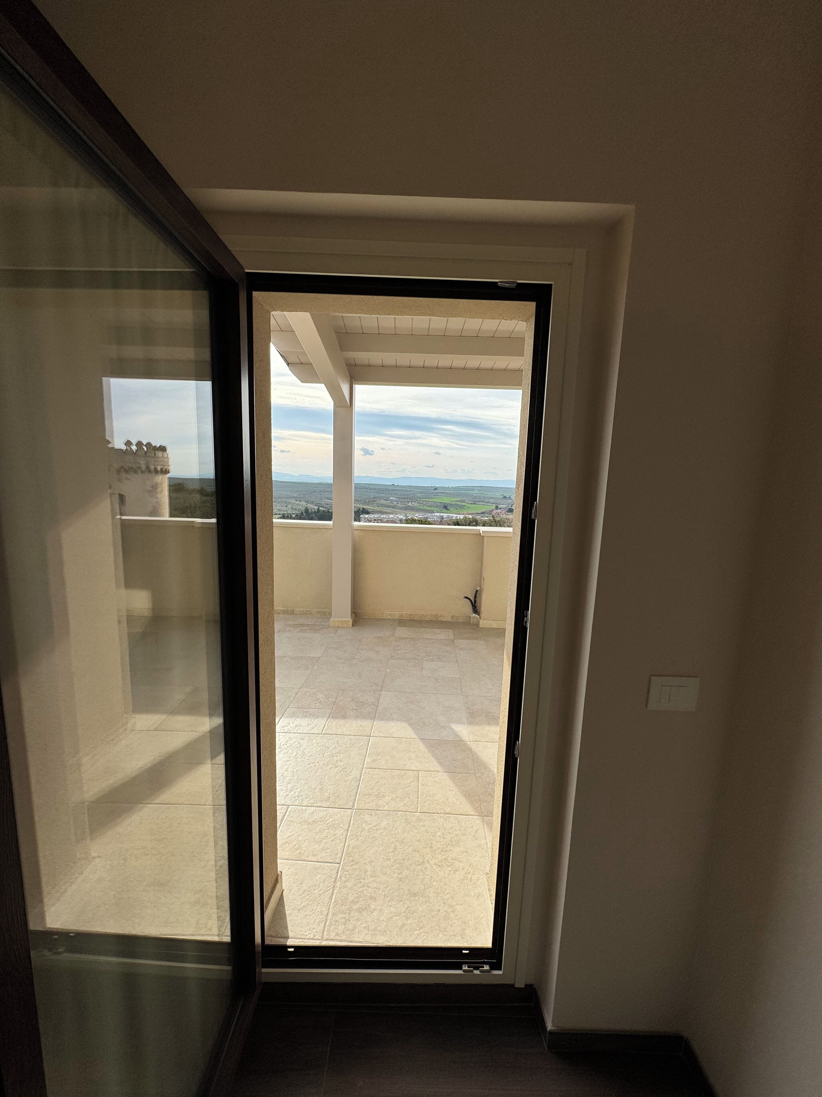 Une pièce dotée d’une porte-fenêtre coulissante donnant sur un balcon carrelé, offrant une vue sur un paysage.