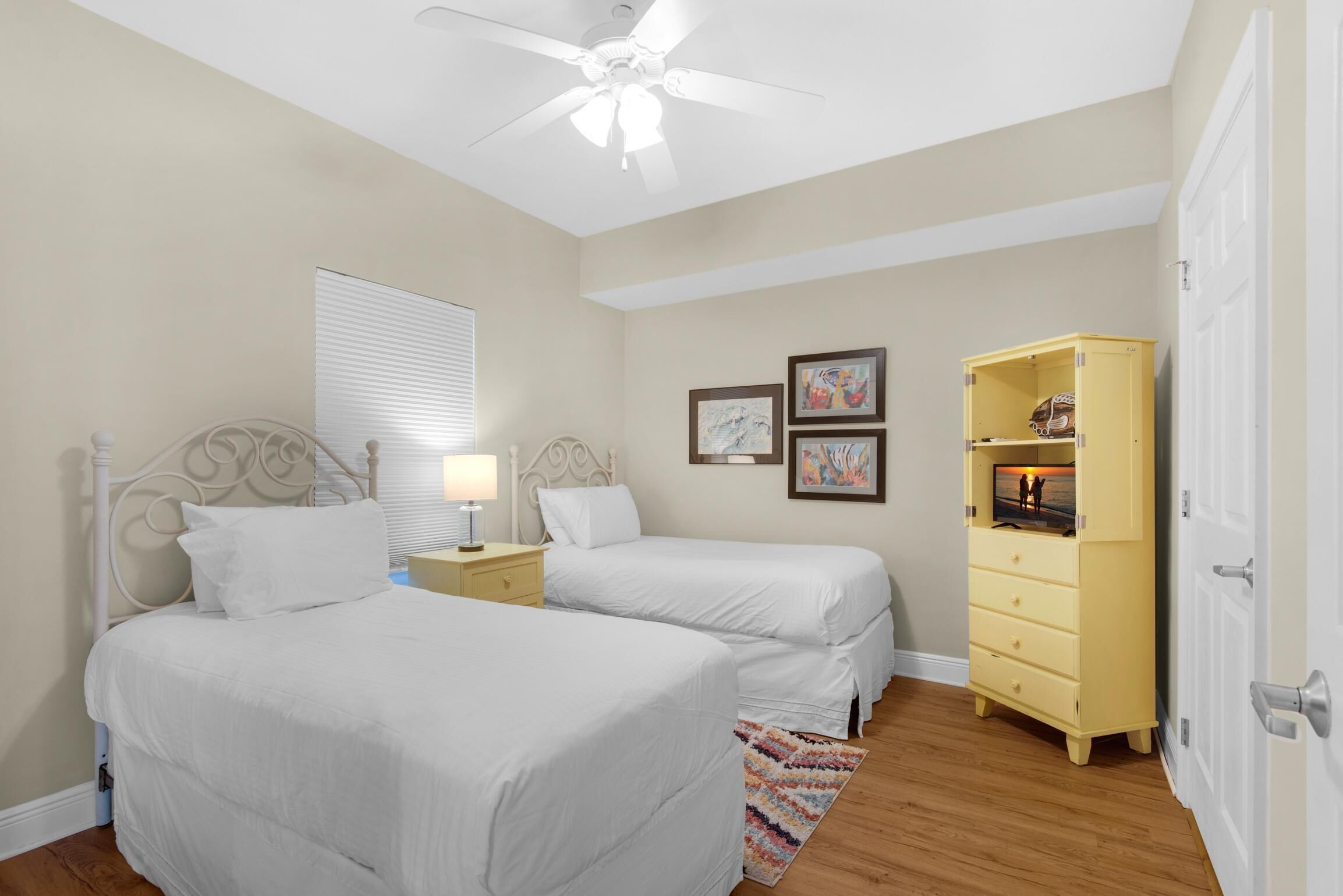 A bedroom with two beds, a ceiling fan, a bedside table with a lamp, a window with blinds, and a yellow dresser.