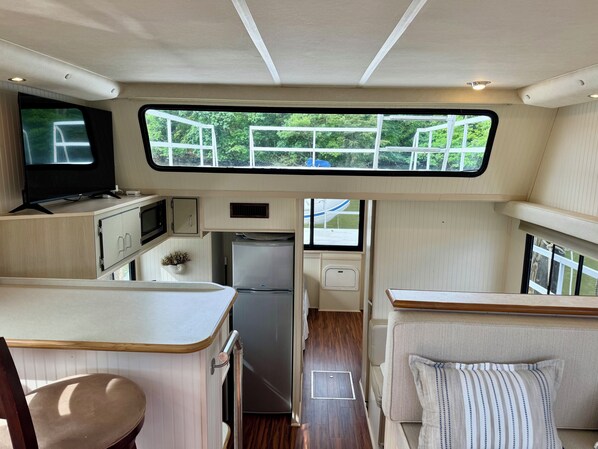 A compact RV interior with a kitchenette, a seating area, and a window overlooking greenery.