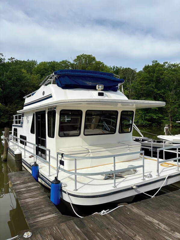 Een wit woonschip afgemeerd aan een houten steiger, met een overdekt dek en een klein huisje.