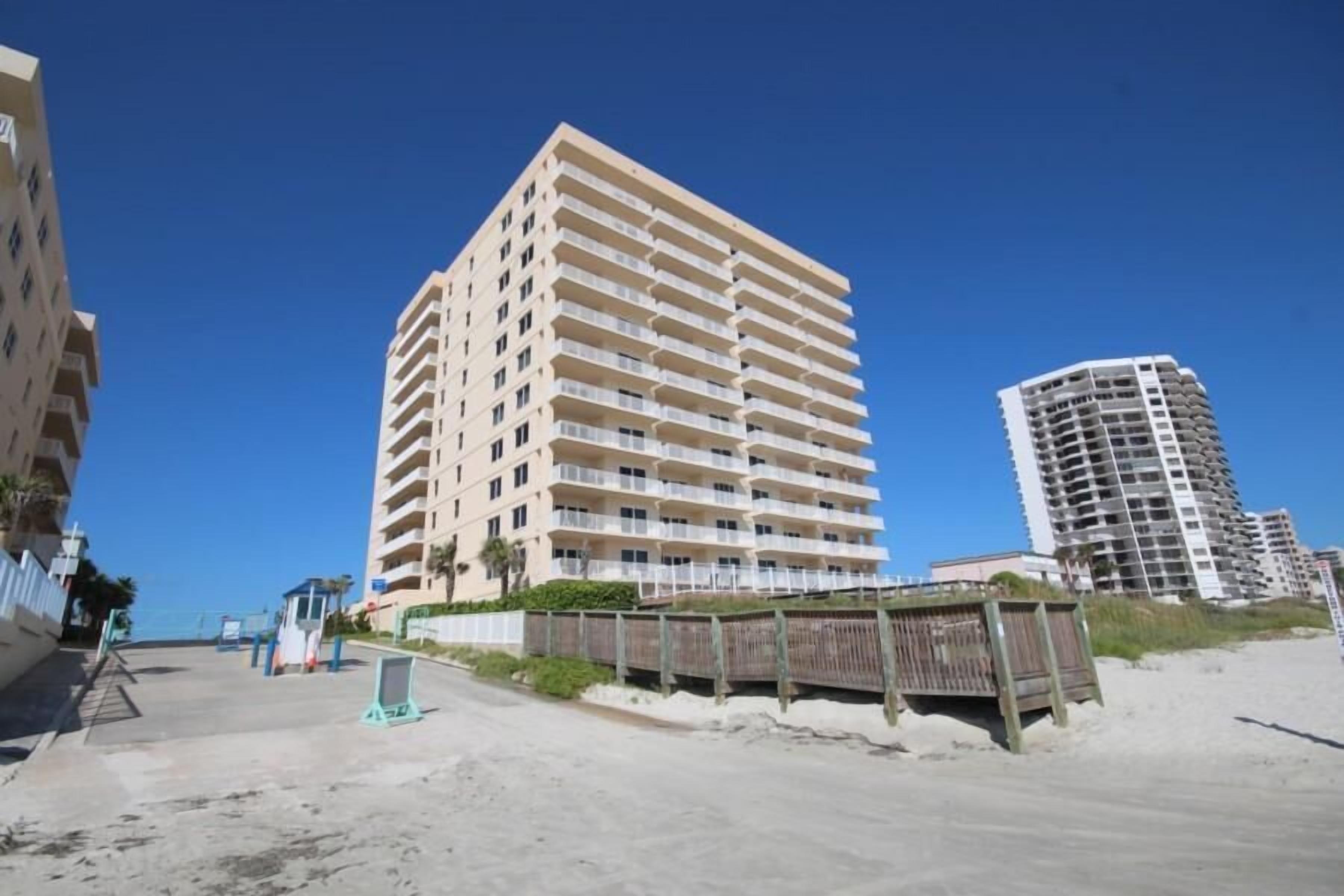 A beachfront hotel with multiple balconies, a sandy beach in the foreground, and other buildings in the background.