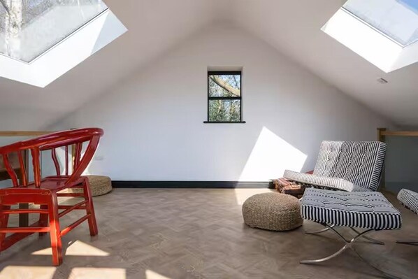 Ein Zimmer mit hohen Decken und geneigter Dachschräge, einem kleinen Fenster und Blick auf Bäume. Zwei rote Stühle und ein gestreifter Hocker sind vorhanden.