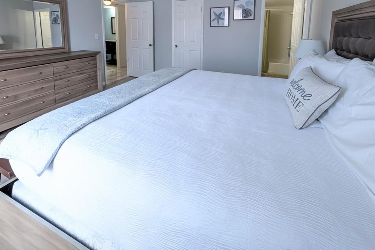 Una cama bien hecha con edredón blanco y una almohada con el texto 'Welcome Home'.