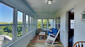 A covered porch with large windows, a rocking chair, a sofa, and a small table. - 703 By The Sea by Topsail Realty (Holly Ridge)