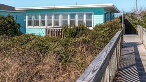 A turquoise building with white-framed windows, surrounded by dense shrubbery and a wooden walkway leading to it. - 703 By The Sea by Topsail Realty (Holly Ridge)